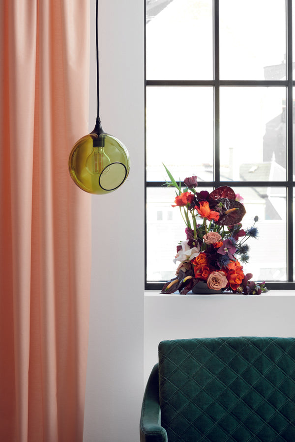 Rund lampe i grøn glas med et afskåret stykke med kig til pæren. Fatning er sort og ledningen er af sort stof.